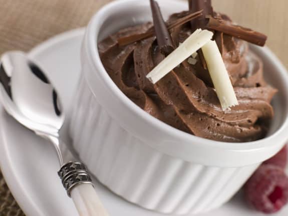 Schokoladenmousse in weißem Schälchen mit Schokospänen, daneben ein Löffel und eine Himbeere.