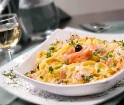 Ein Teller Pasta mit Lachs und Kräutern, garniert mit einer Olive, neben einem Glas Weißwein.