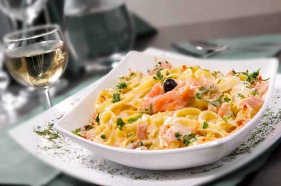 Ein Teller Pasta mit Lachs und Kräutern, garniert mit einer Olive, neben einem Glas Weißwein.