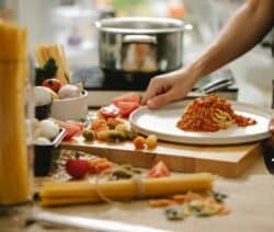 Spaghetti mit Tomatensauce wird auf einem Teller serviert, umgeben von rohen Zutaten und einem Kochtopf im Hintergrund.