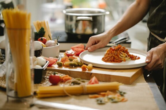 Spaghetti mit Tomatensauce wird auf einem Teller serviert, umgeben von rohen Zutaten und einem Kochtopf im Hintergrund.