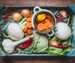 Gemüsebox mit Karotten, Zwiebeln, Knoblauch, Chili, Kohlrabi und einem Topf mit geschnittenen Karotten auf Holztisch.