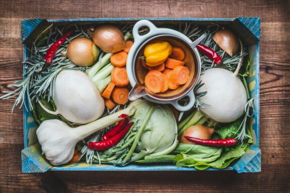 Gemüsebox mit Karotten, Zwiebeln, Knoblauch, Chili, Kohlrabi und einem Topf mit geschnittenen Karotten auf Holztisch.
