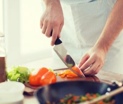 Person schneidet orange Paprika auf einem Holzbrett in der Küche, daneben Salat, Ölflasche und Pfanne.