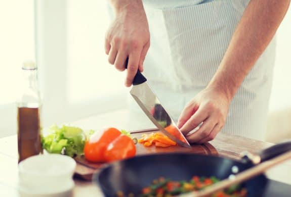 Person schneidet orange Paprika auf einem Holzbrett in der Küche, daneben Salat, Ölflasche und Pfanne.