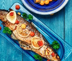 Ein gebratener Fisch mit Zitronenscheiben und Tomaten auf blauem Teller, daneben gelbe Tomaten in einer Schüssel.