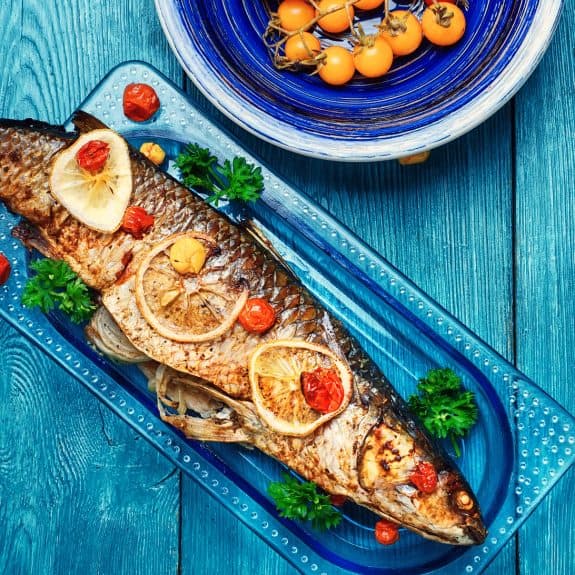 Ein gebratener Fisch mit Zitronenscheiben und Tomaten auf blauem Teller, daneben gelbe Tomaten in einer Schüssel.