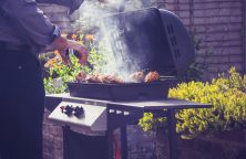 Eine Person grillt Fleisch auf einem rauchenden Grill im Garten, umgeben von Blumen und Sträuchern.