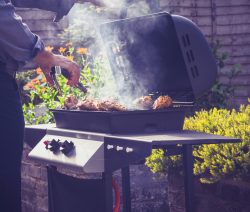 Eine Person grillt Fleisch auf einem rauchenden Grill im Garten, umgeben von Blumen und Sträuchern.