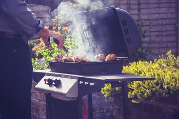 Eine Person grillt Fleisch auf einem rauchenden Grill im Garten, umgeben von Blumen und Sträuchern.