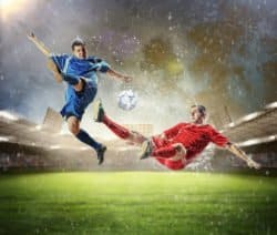 Zwei Fußballspieler in Blau und Rot kämpfen in der Luft um den Ball im Stadion bei Regen.