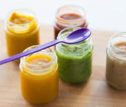 Fünf Gläser mit buntem Püree, lila Löffel auf grünem Glas, auf Holzuntergrund.
