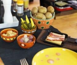 Bunter Tisch mit Raclette-Pfännchen, Kartoffeln, Gurken, Perlzwiebeln und Speck auf einem dunklen Tuch.
