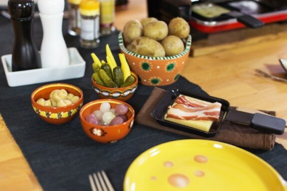 Bunter Tisch mit Raclette-Pfännchen, Kartoffeln, Gurken, Perlzwiebeln und Speck auf einem dunklen Tuch.