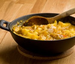 Ein schwarzer Topf mit Kartoffelcurry und Holzlöffel auf einem Holztisch.
