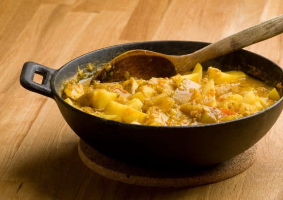 Ein schwarzer Topf mit Kartoffelcurry und Holzlöffel auf einem Holztisch.