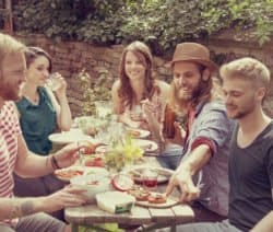Fünf Freunde sitzen im Garten um einen Tisch mit Essen und Getränken, lächelnd und plaudernd.