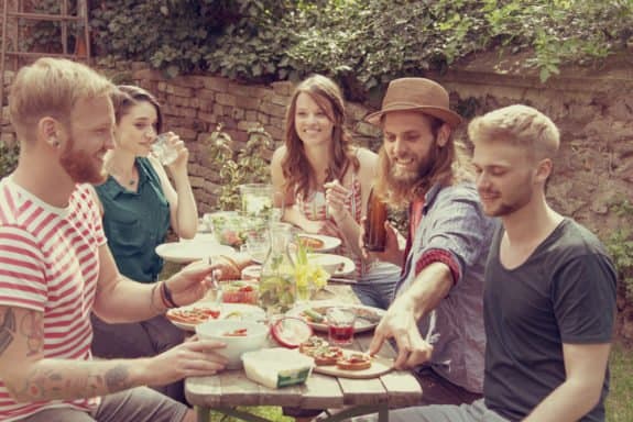 Fünf Freunde sitzen im Garten um einen Tisch mit Essen und Getränken, lächelnd und plaudernd.