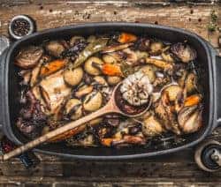 Ein Bräter mit gebratenem Gemüse und Kräutern auf einem Holztisch, umgeben von Gewürzen und Kräutern.