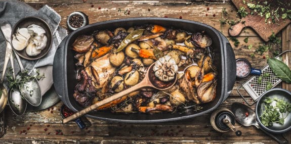 Ein Bräter mit gebratenem Gemüse und Kräutern auf einem Holztisch, umgeben von Gewürzen und Kräutern.