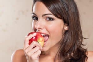 Eine Frau beißt in einen roten Apfel, lächelt und schaut in die Kamera.