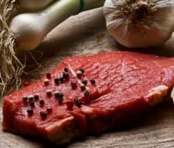 Ein rohes Steak mit Pfefferkörnern liegt auf Holz, umgeben von Knoblauch, Frühlingszwiebeln und einer Zitronenscheibe.