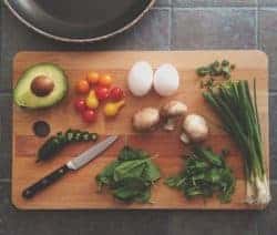 Avocado, Tomaten, Eier, Pilze, Frühlingszwiebeln und Spinat auf einem Holzbrett mit Messer. Pfanne im Hintergrund.