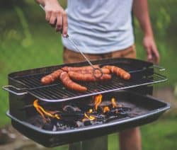 Person grillt Würste auf einem Holzkohlegrill, Flammen lodern darunter.