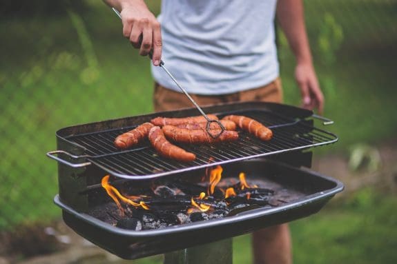 Person grillt Würste auf einem Holzkohlegrill, Flammen lodern darunter.