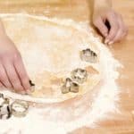 Hände schneiden Plätzchenformen aus ausgerolltem Teig auf bemehltem Holztisch.