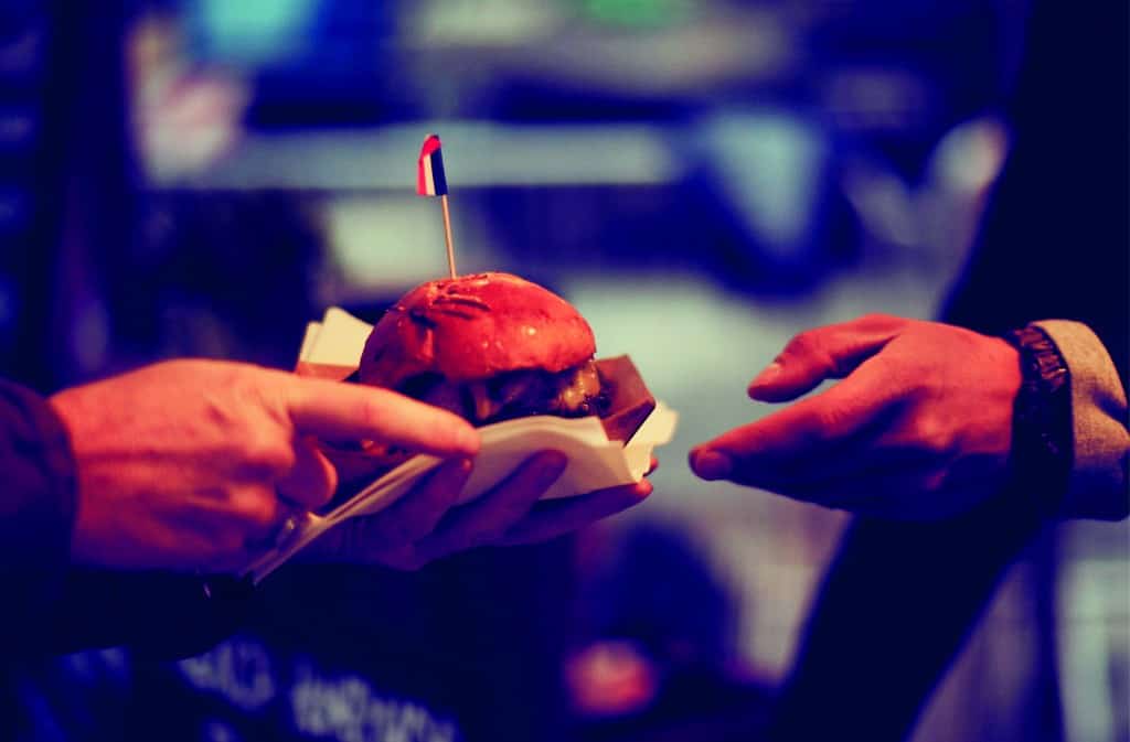 Ein Burger mit Zahnstocherflagge wird zwischen zwei Händen übergeben.