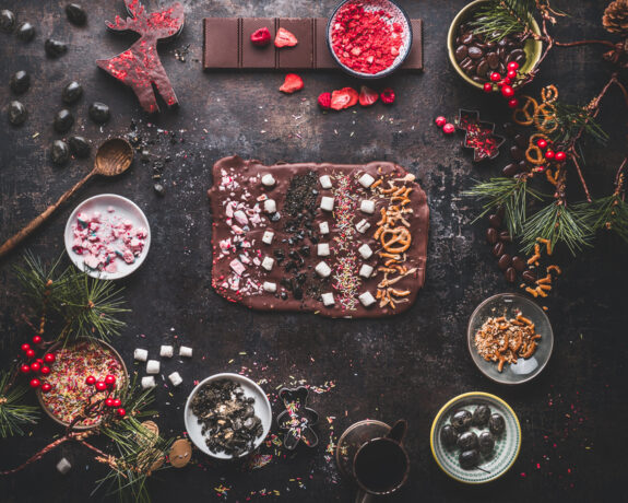 Selbstgemachte Schokolade mit Nüssen und Beeren, umgeben von weihnachtlicher Dekoration und Zutaten auf dunklem Tisch.