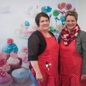 Zwei Frauen in roten Schürzen vor einem Hintergrund mit bunten Cupcakes.