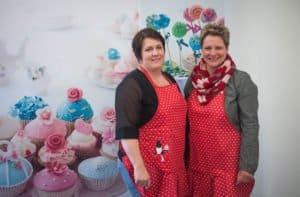 Zwei Frauen in roten Schürzen vor einem Hintergrund mit bunten Cupcakes.