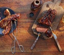 Gegrilltes Fleisch auf Holzbrett und Teller, mit Grillzange, Barbecuesoße und Glas auf Holztisch.