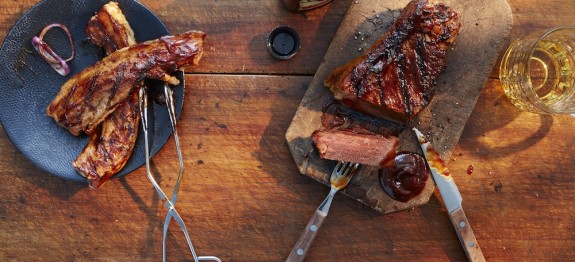 Gegrilltes Fleisch auf Holzbrett und Teller, mit Grillzange, Barbecuesoße und Glas auf Holztisch.