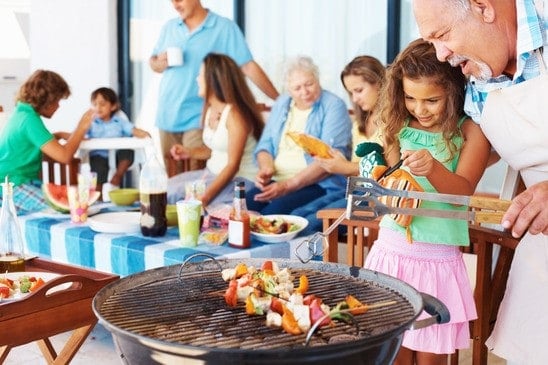 Eine Familie grillt im Freien; ein Mann und ein Mädchen wenden Spieße, während andere am Tisch essen.