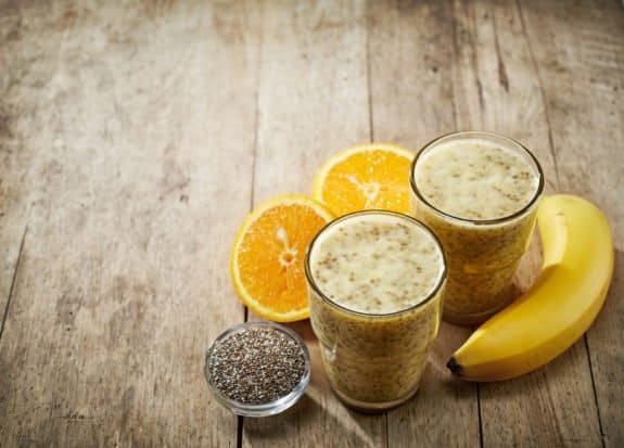 Zwei Gläser Smoothie, Banane, Orangenscheiben und Chiasamen auf Holztisch.
