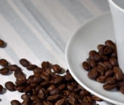 Weiße Tasse auf Untertasse, umgeben von verstreuten Kaffeebohnen auf gestreifter Tischdecke.