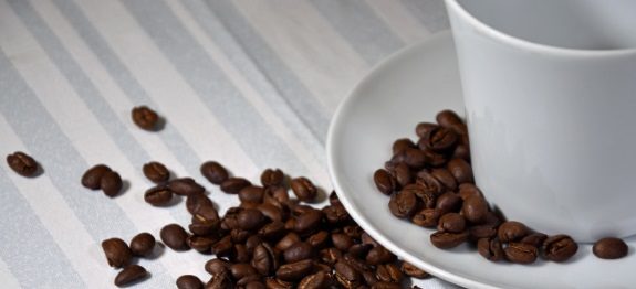 Weiße Tasse auf Untertasse, umgeben von verstreuten Kaffeebohnen auf gestreifter Tischdecke.