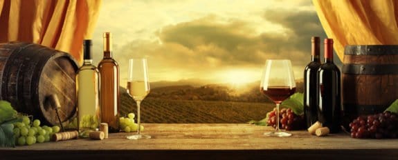 Weinflaschen und Gläser auf einem Tisch mit Trauben, Fässern und Sonnenuntergang über Weinbergen im Hintergrund.