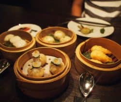 Verschiedene Dim Sum in Bambuskörben auf einem Tisch, daneben ein Löffel und ein Teller.