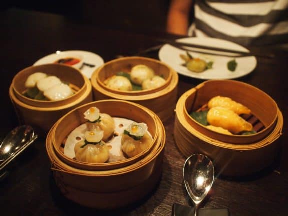 Verschiedene Dim Sum in Bambuskörben auf einem Tisch, daneben ein Löffel und ein Teller.