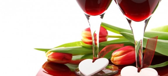 Zwei Weingläser mit Herzen dekoriert, umgeben von roten Tulpen auf glänzendem Tisch.