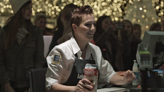 Eine Kassiererin in Edeka-Uniform scannt Kekse, umgeben von Menschen und Lichtern im Hintergrund.