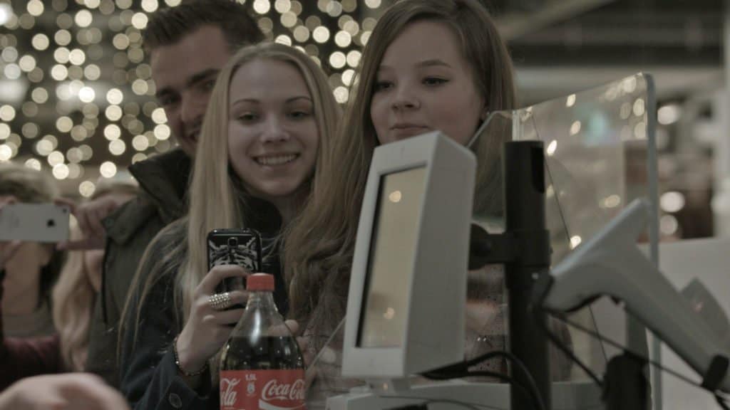 Zwei lachende junge Frauen machen ein Selfie vor einem beleuchteten Hintergrund mit einer Coca-Cola-Flasche im Vordergrund.