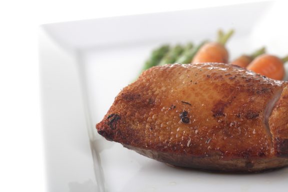 Gebratenes Entenbrustfilet auf weißem Teller, im Hintergrund Spargel und Karotten.