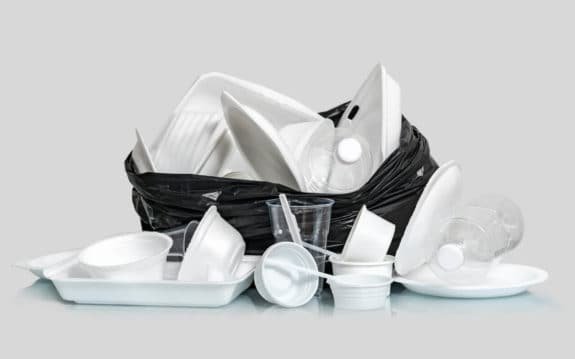 Ein schwarzer Müllsack voller Einwegplastikgeschirr und -flaschen auf grauem Hintergrund.