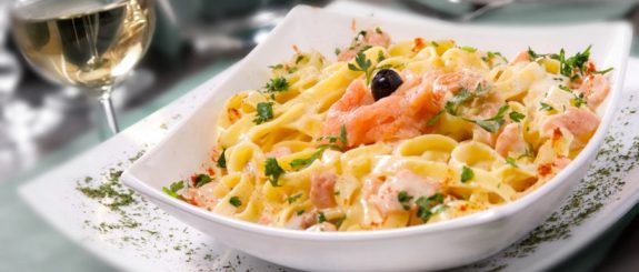 Dekorativer Teller mit Lachs Bandnudeln und einem Glas Wein im Hintergrund