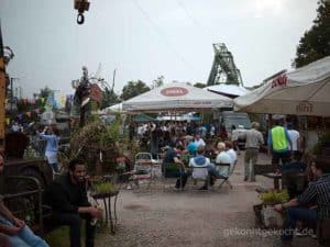 Ein belebter Biergarten mit Menschen unter Sonnenschirmen, umgeben von Pflanzen und Dekorationen. Im Hintergrund ein Förderturm.